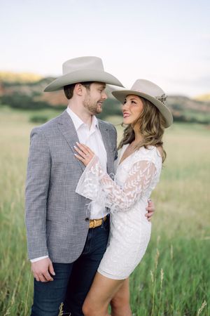 A romantic couple in cowboy hats embrace in a sunlit field wearing western attire and formal clothing.