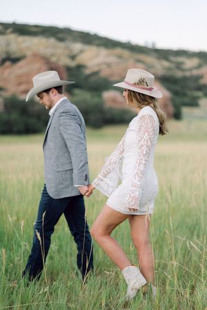 A couple in western attire playfully walks through a grassy meadow during a countryside photoshoot.