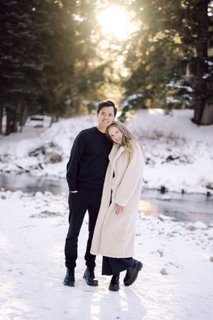 A couple shares a romantic moment in a snowy winter forest setting with sunlight filtering through the trees.