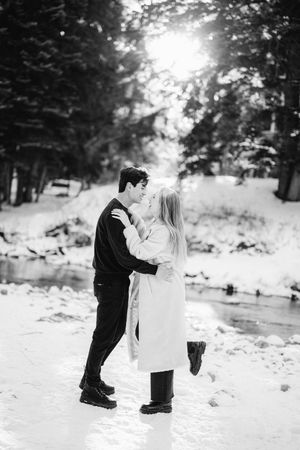 A couple shares a romantic moment in the snow surrounded by evergreen trees and winter scenery.