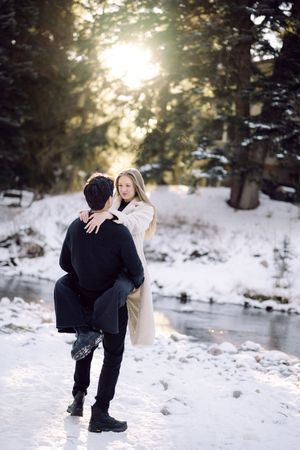 A couple shares a romantic moment in a snowy winter landscape with sunlight filtering through evergreen trees.