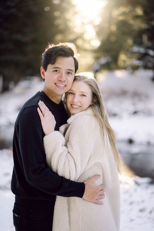 A couple embraces outdoors in a snowy winter setting with sunlit trees in the background.