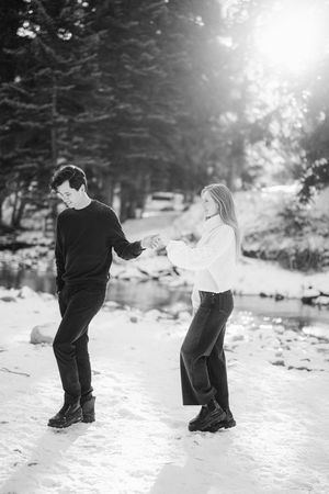 A couple playfully dances together in a snowy winter forest setting captured in black and white photography.