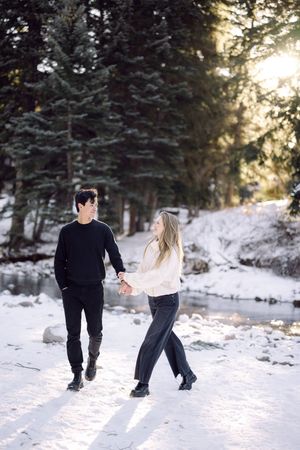 A couple plays together in a snowy winter forest scene with sunlight filtering through evergreen trees.
