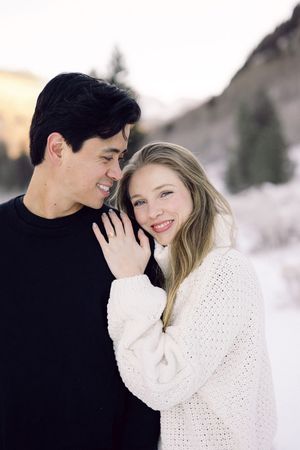 A romantic winter engagement photo shows a couple in black and white sweaters embracing in the snow.
