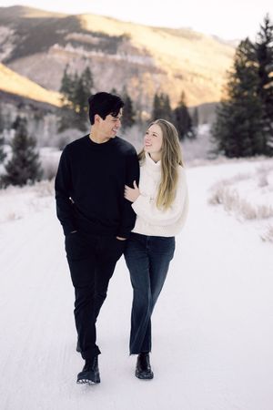 Couple walking together in a snowy winter landscape with mountains and evergreen trees in the background.