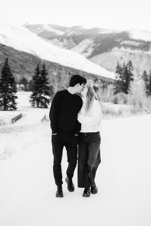 A romantic black and white winter scene shows a couple sharing an intimate moment while walking in the snowy mountains.