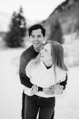 A couple embraces and laughs together in a snowy winter setting wearing casual clothing in black and white photography.