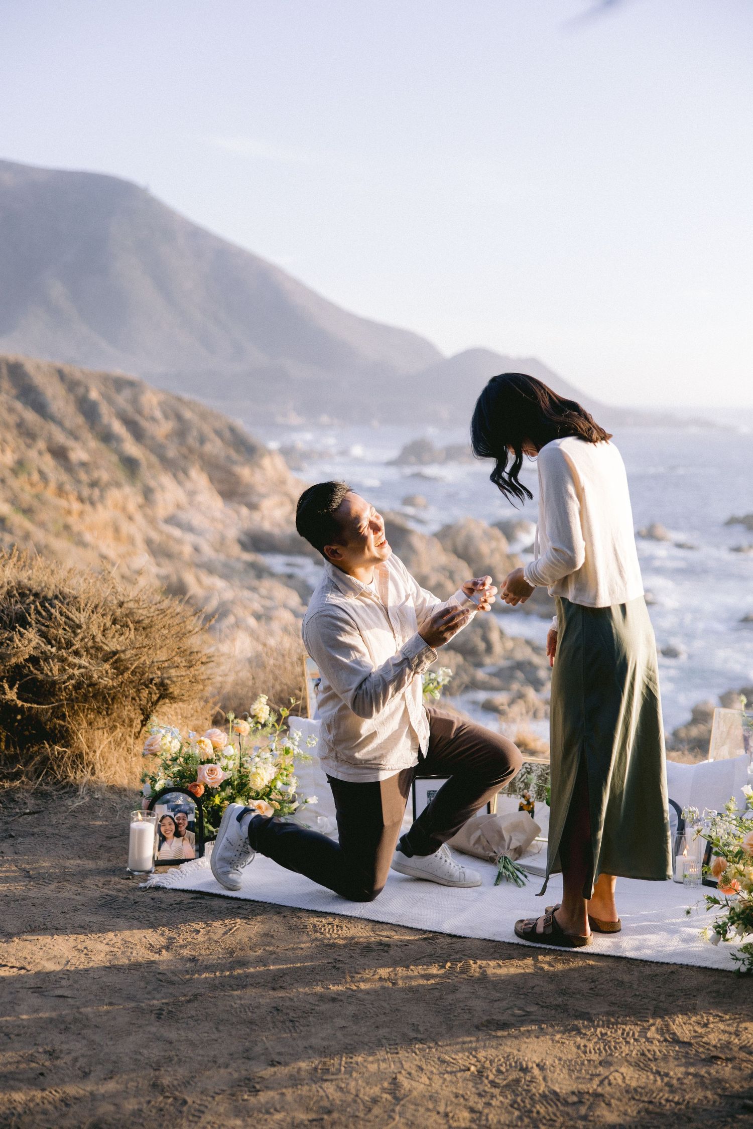 A romantic proposal takes place on a scenic cliff overlooking the ocean with flowers and a white picnic setup at sunset.