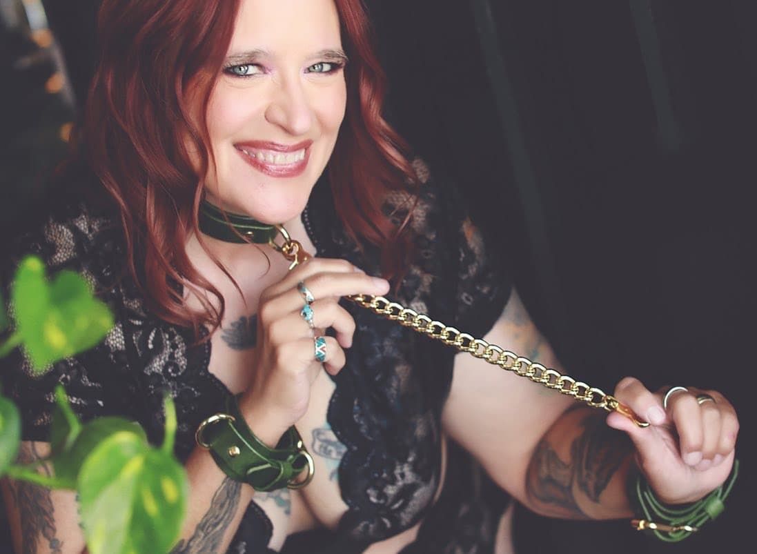 Person with red curly hair holds a chain while wearing black lace and smiling against a blurred green background.