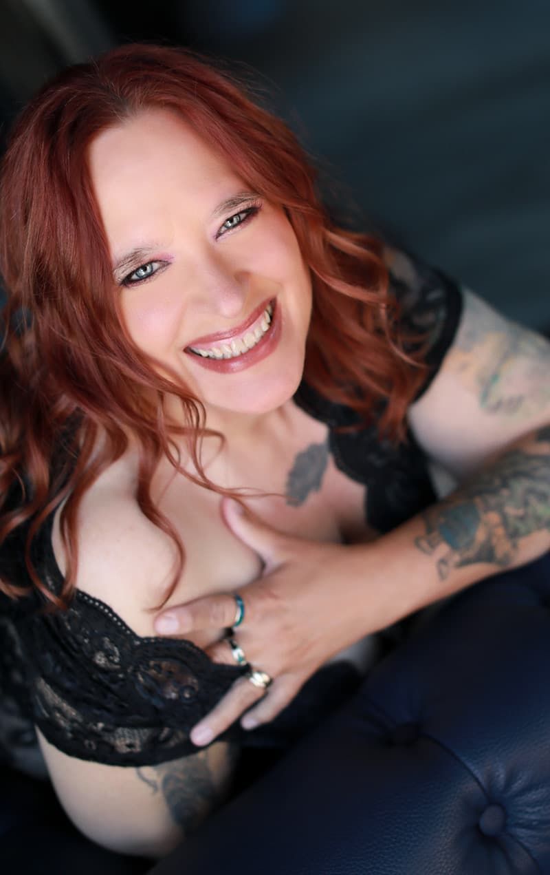 A smiling woman with red hair and tattoos poses in black lace lingerie against a dark background.