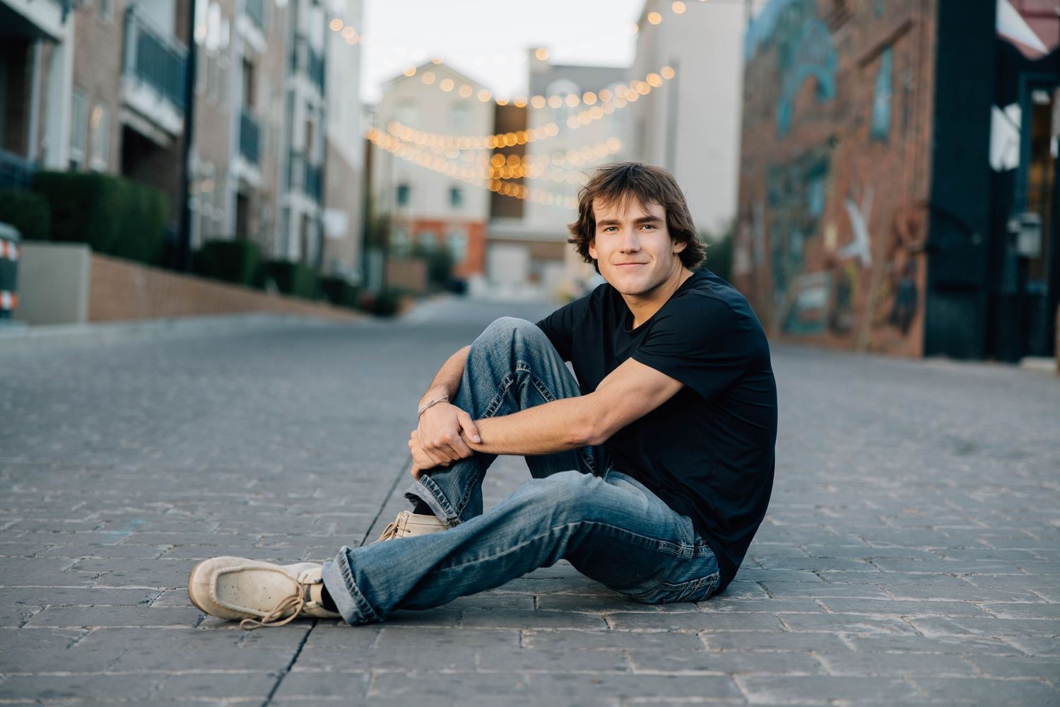 Young man in black t-shirt and jeans sitting casually on urban cobblestone street with string lights behind him.