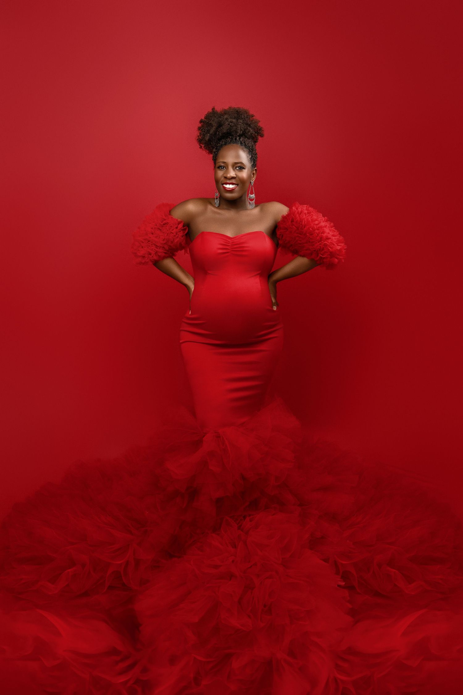 A pregnant woman poses in stunning red mermaid gown with feathered sleeves against vibrant red backdrop.