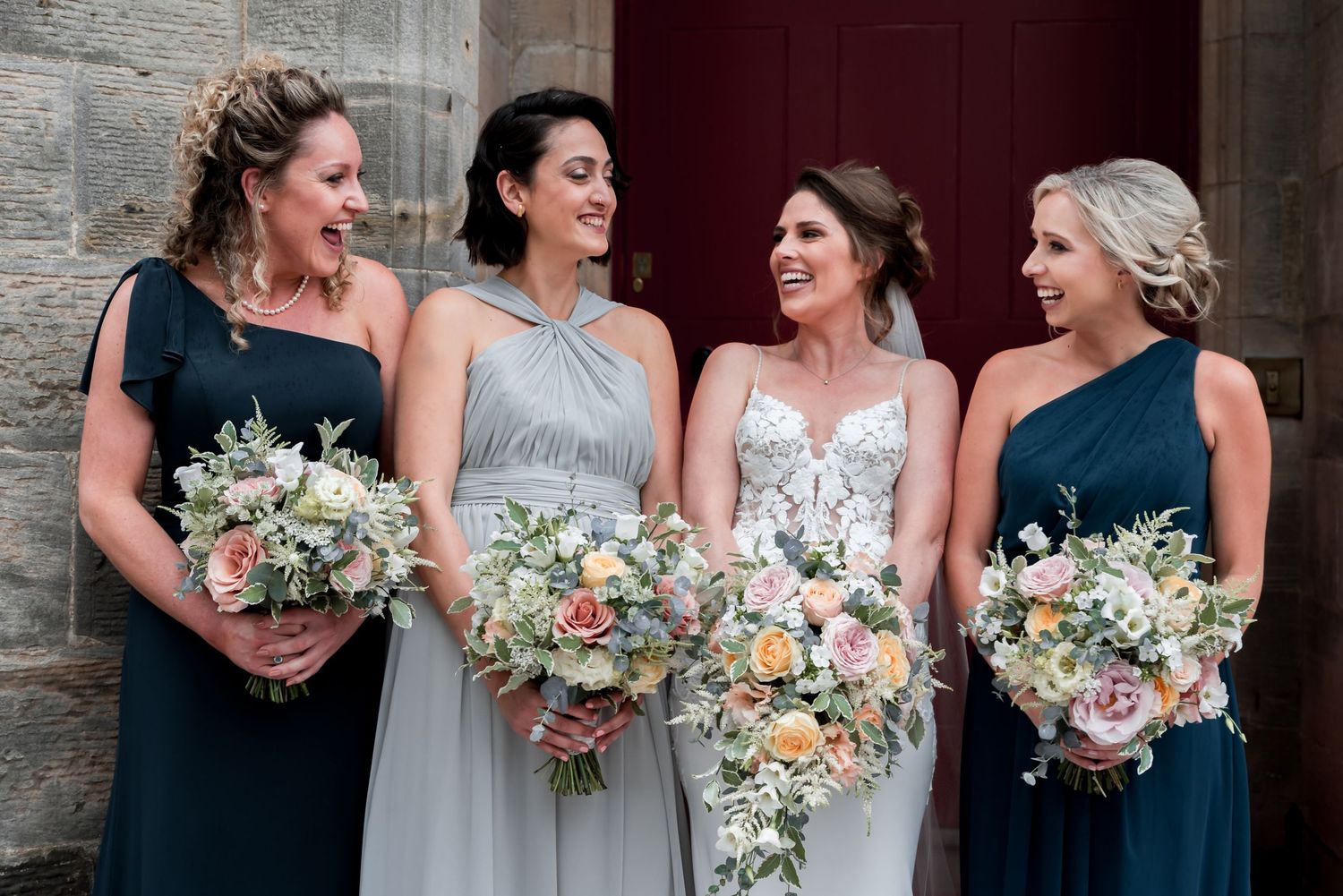 A bridal party poses together holding elegant bouquets with white and peach flowers, wearing navy and silver dresses.
