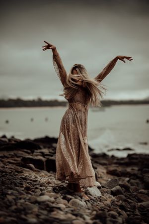 Woman in flowy dress dances on rocky shore with arms raised against moody sky at dusk.