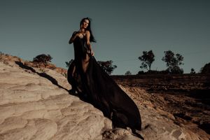 A figure in a flowing black dress poses dramatically on rocky desert terrain at sunset.