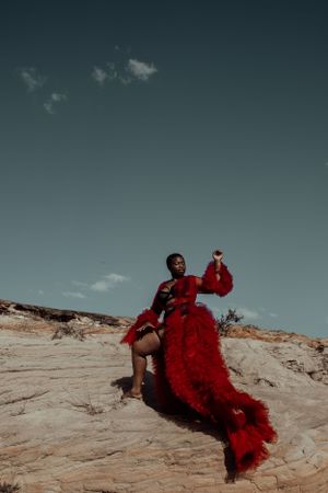 Figure in flowing red gown poses dramatically on desert rock formations against a moody teal sky.