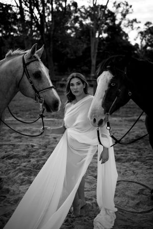 Graceful pose between two horses wearing flowing white dress in monochrome.