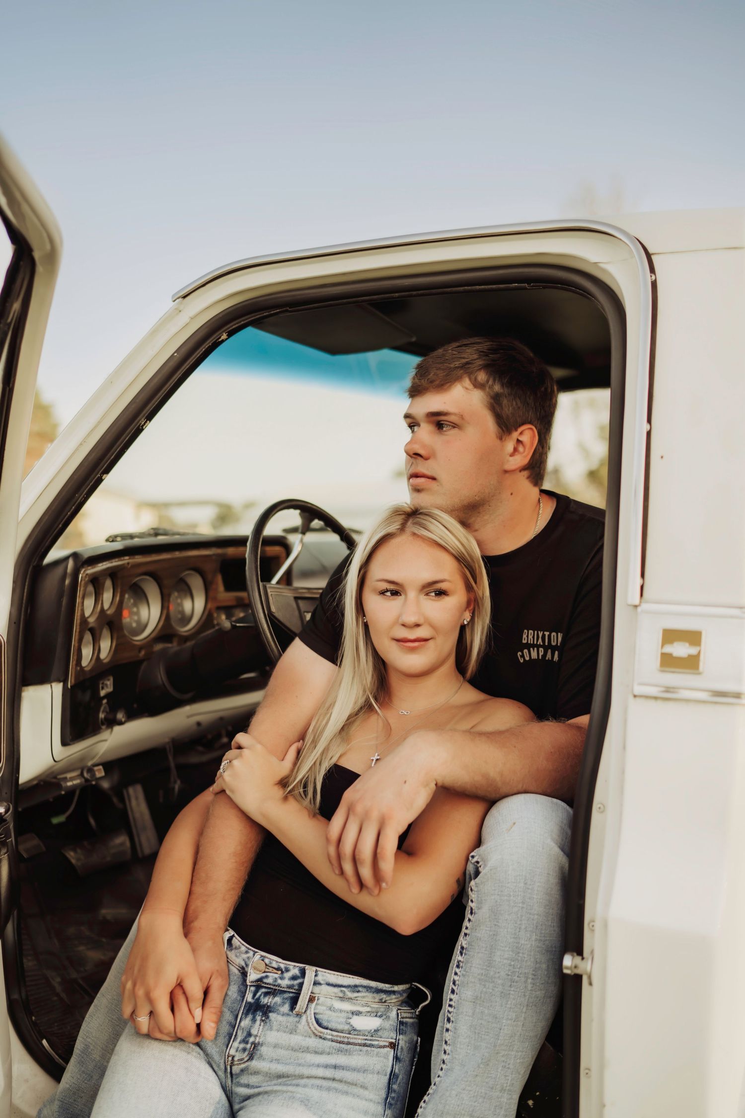 Two people pose together in the open door of a vintage white pickup truck on a sunny day.