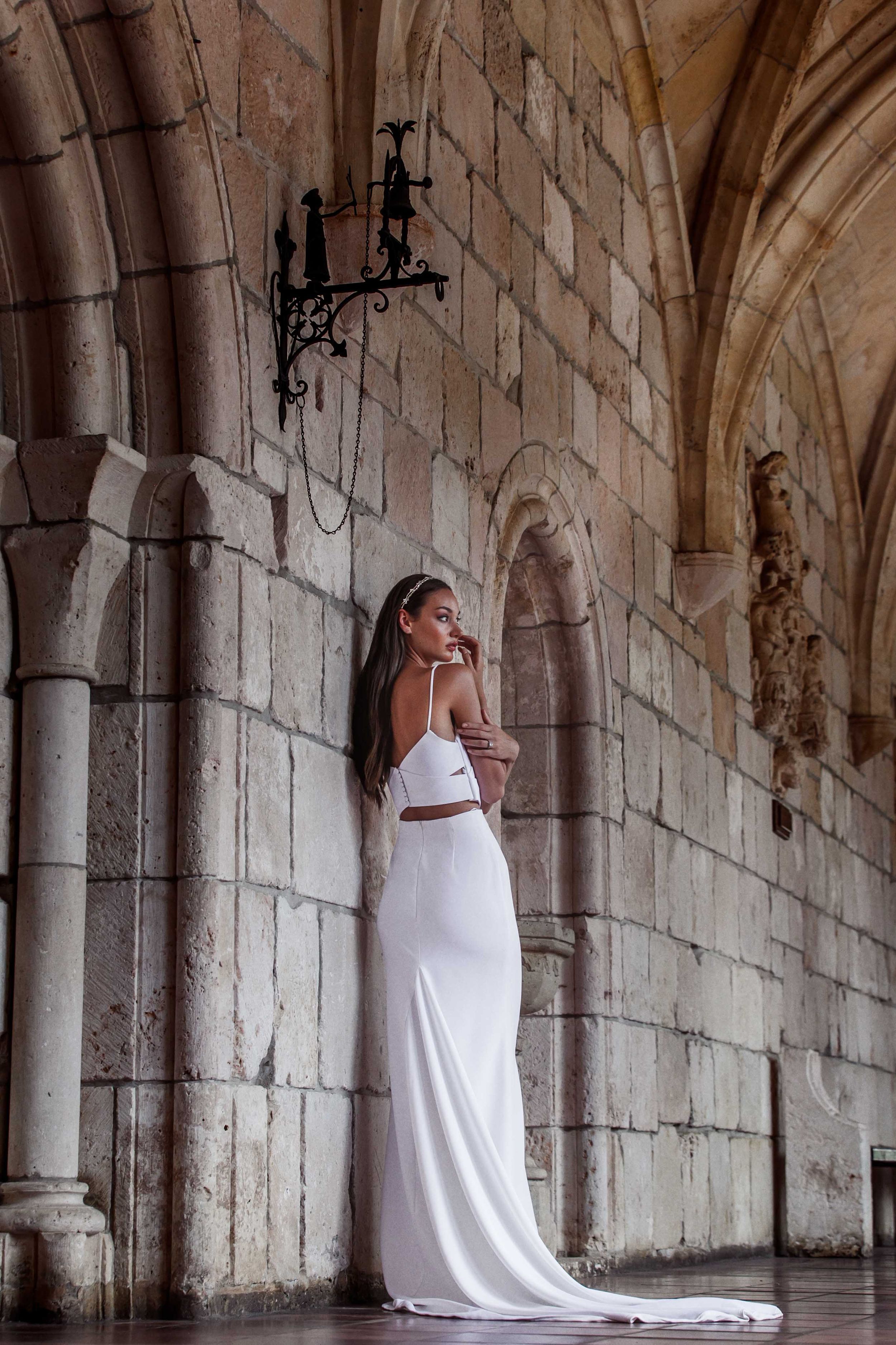 Figure in elegant white flowing dress poses gracefully against stone archways in a historic architectural setting.