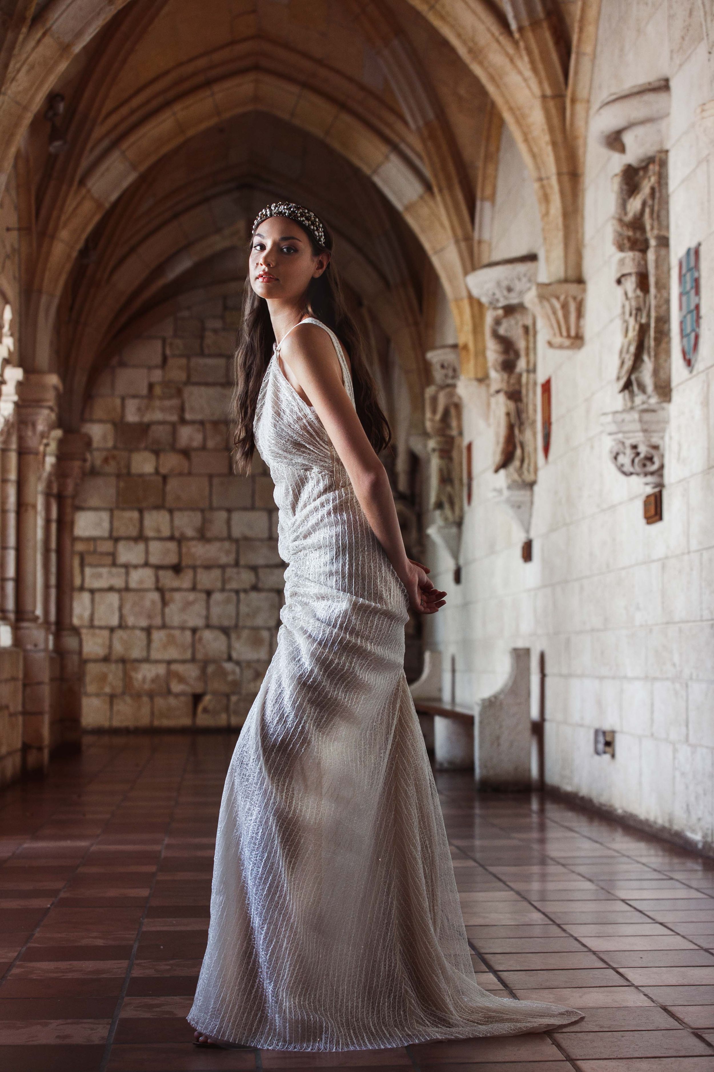 Elegant figure in a flowing white gown poses in a medieval stone archway corridor with ornate architectural details.