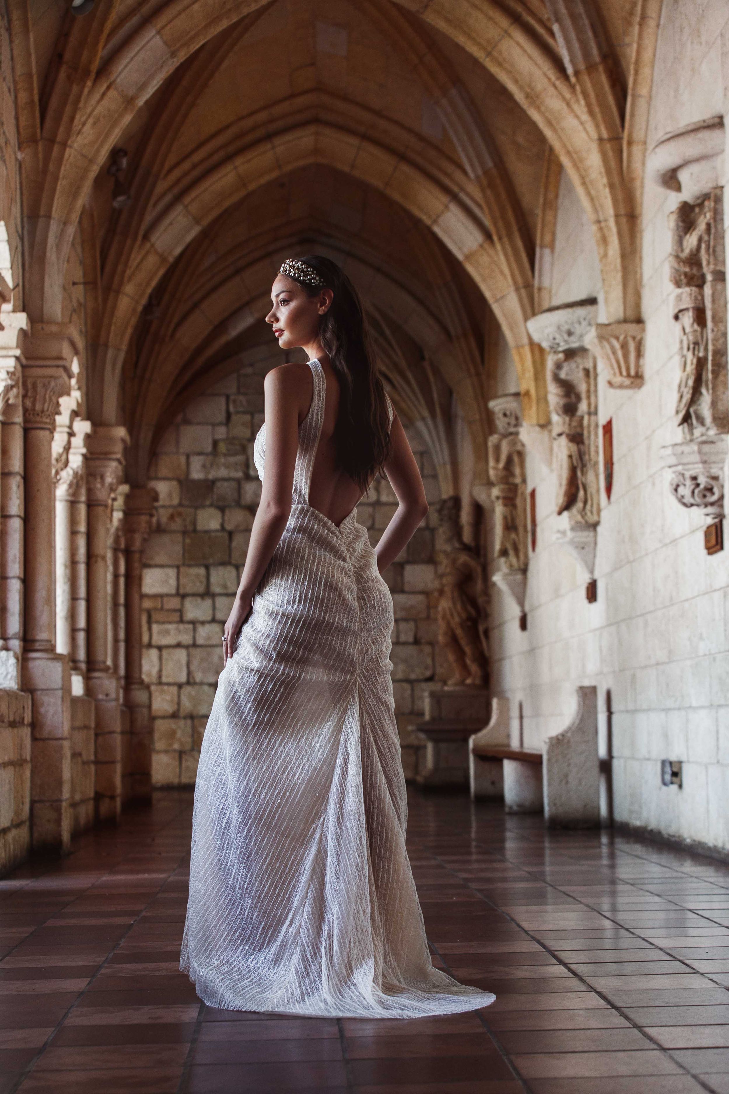 A bride in a white lace dress poses gracefully in a historic stone archway corridor with vaulted ceilings.