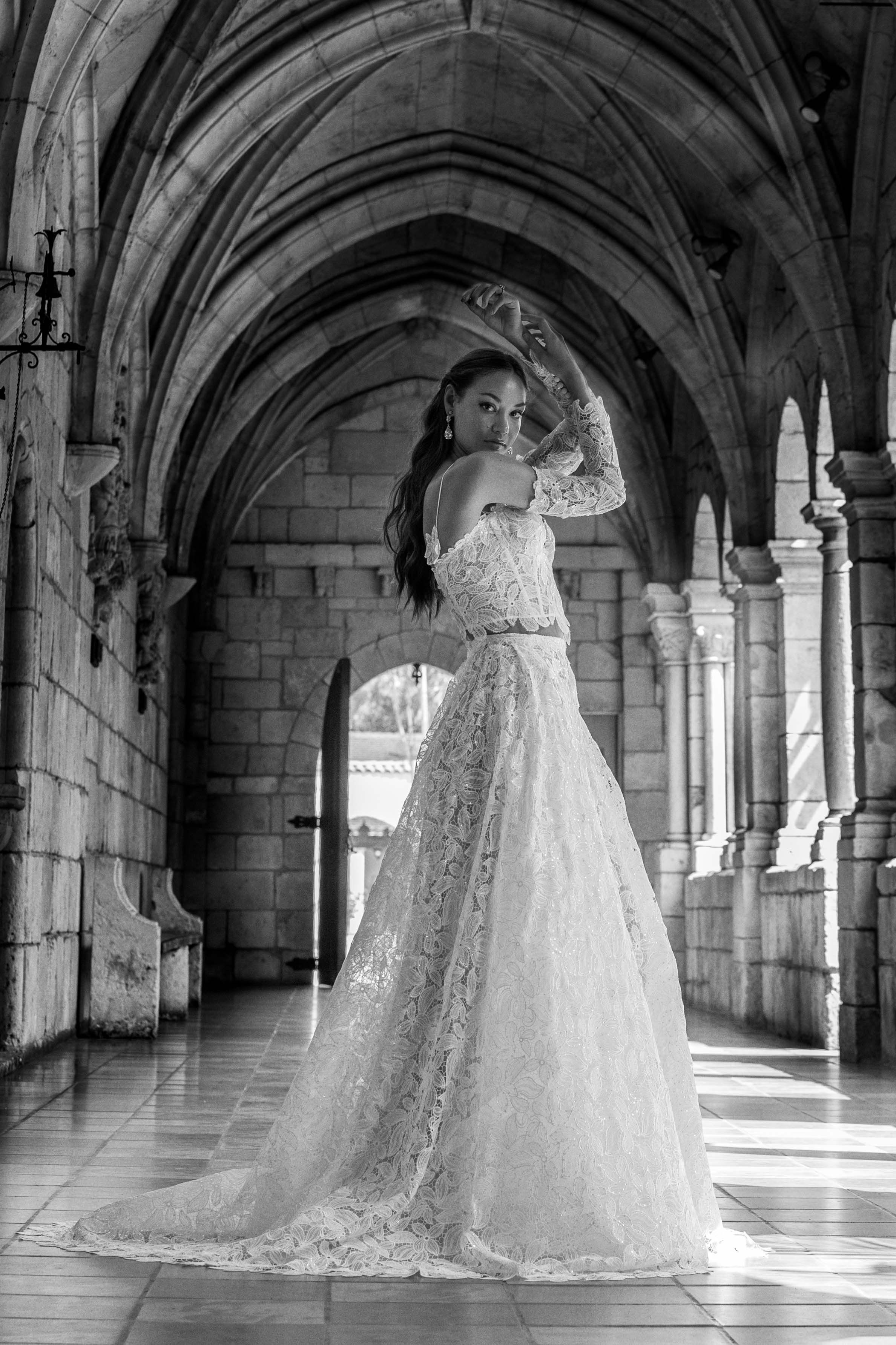 A bride in a flowing lace wedding gown poses gracefully under Gothic stone arches in black and white.