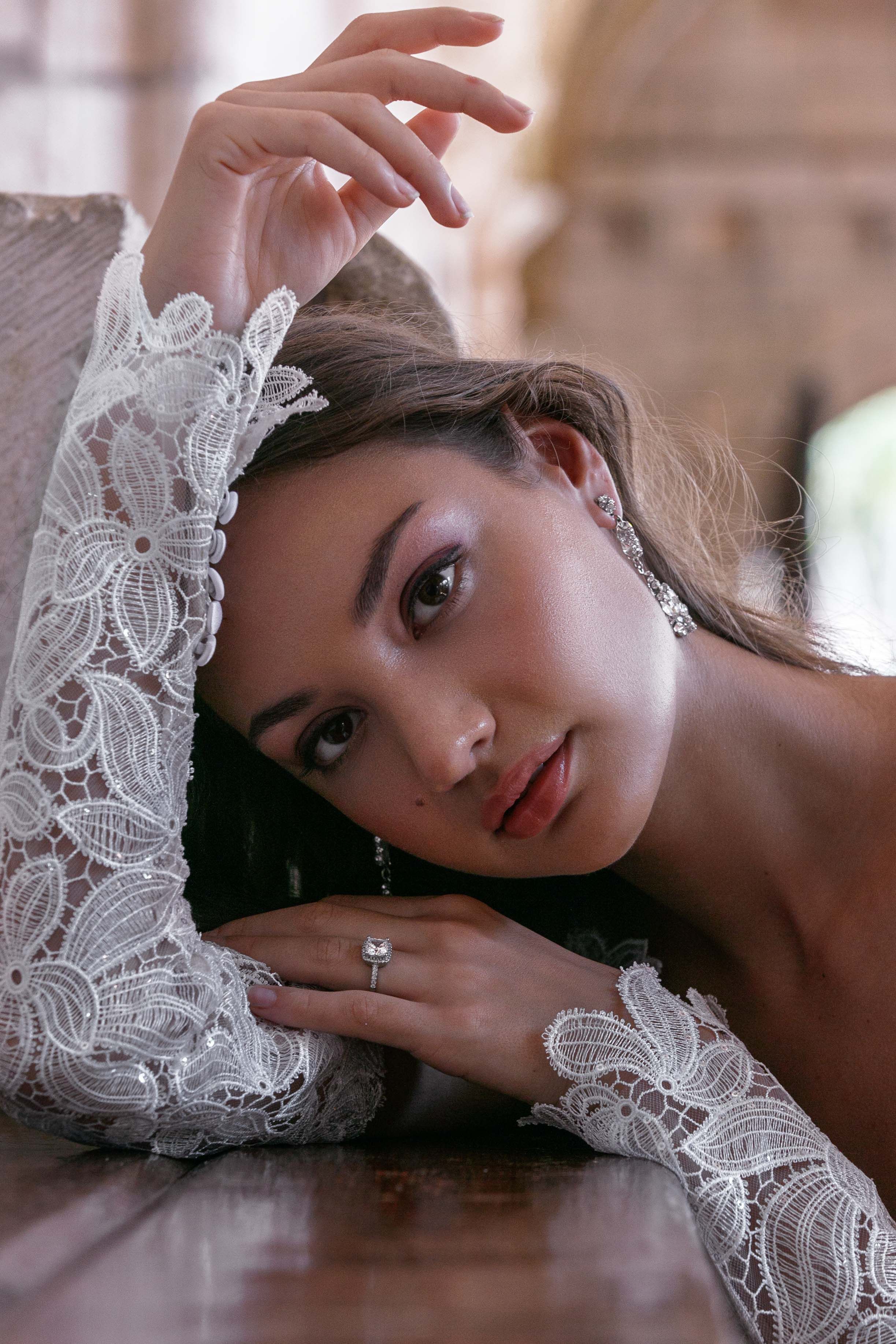 A close-up portrait featuring delicate white lace sleeves, sparkling earrings and a ring in soft romantic lighting.