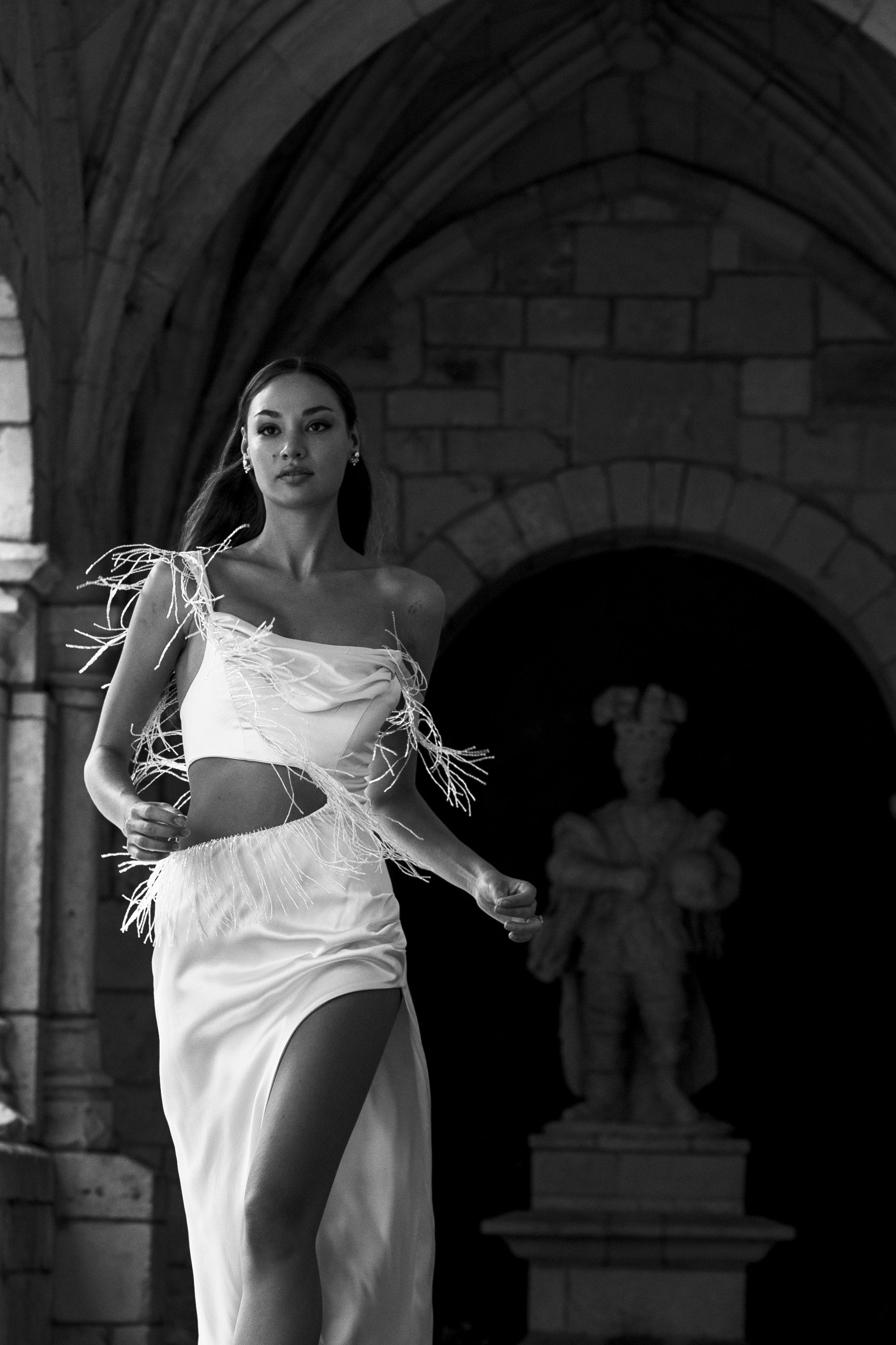 A dancer in a flowing white dress performs dynamic movements in front of a stone archway in black and white.