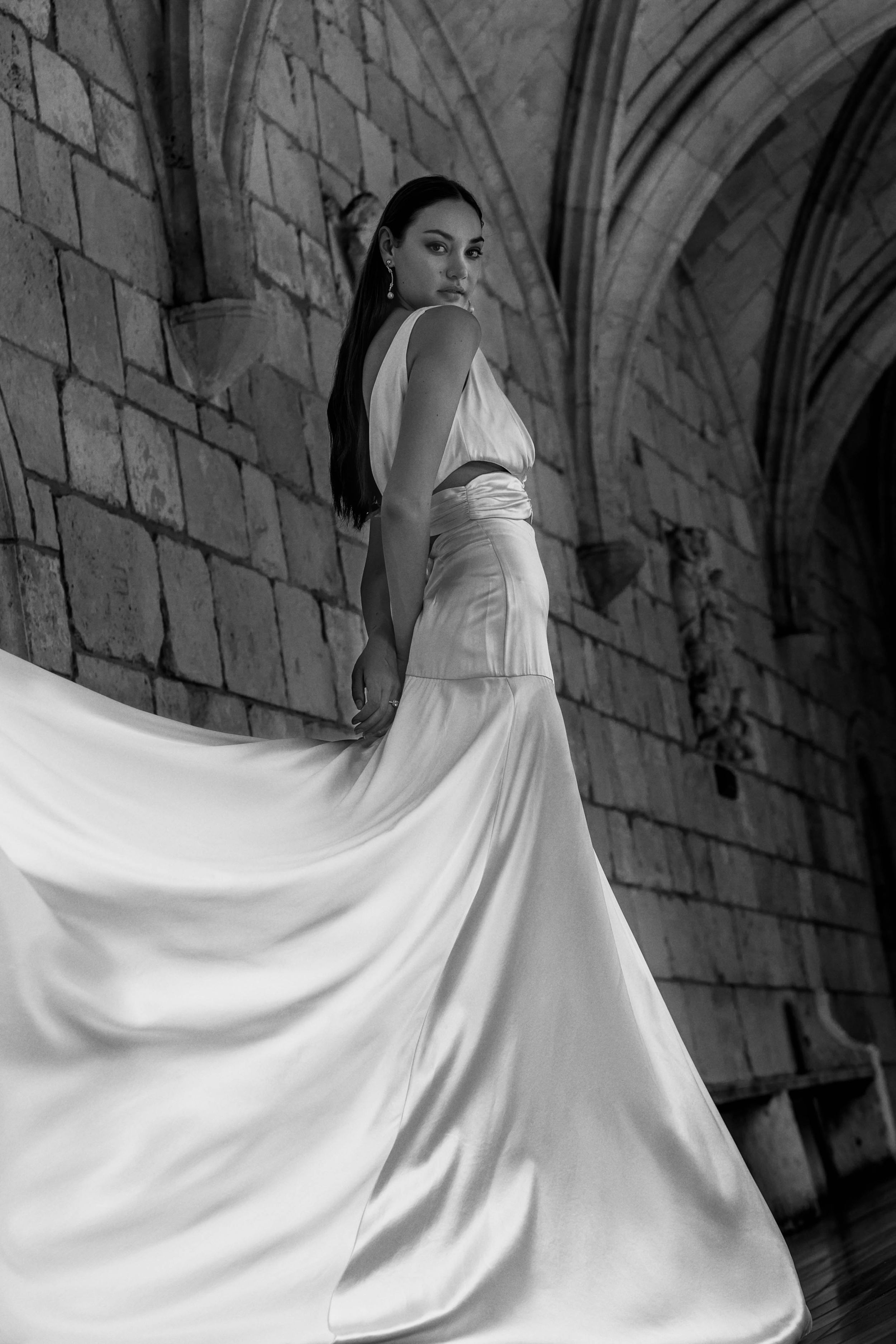 A person in a flowing white wedding gown poses elegantly in a Gothic stone corridor with arched architecture.