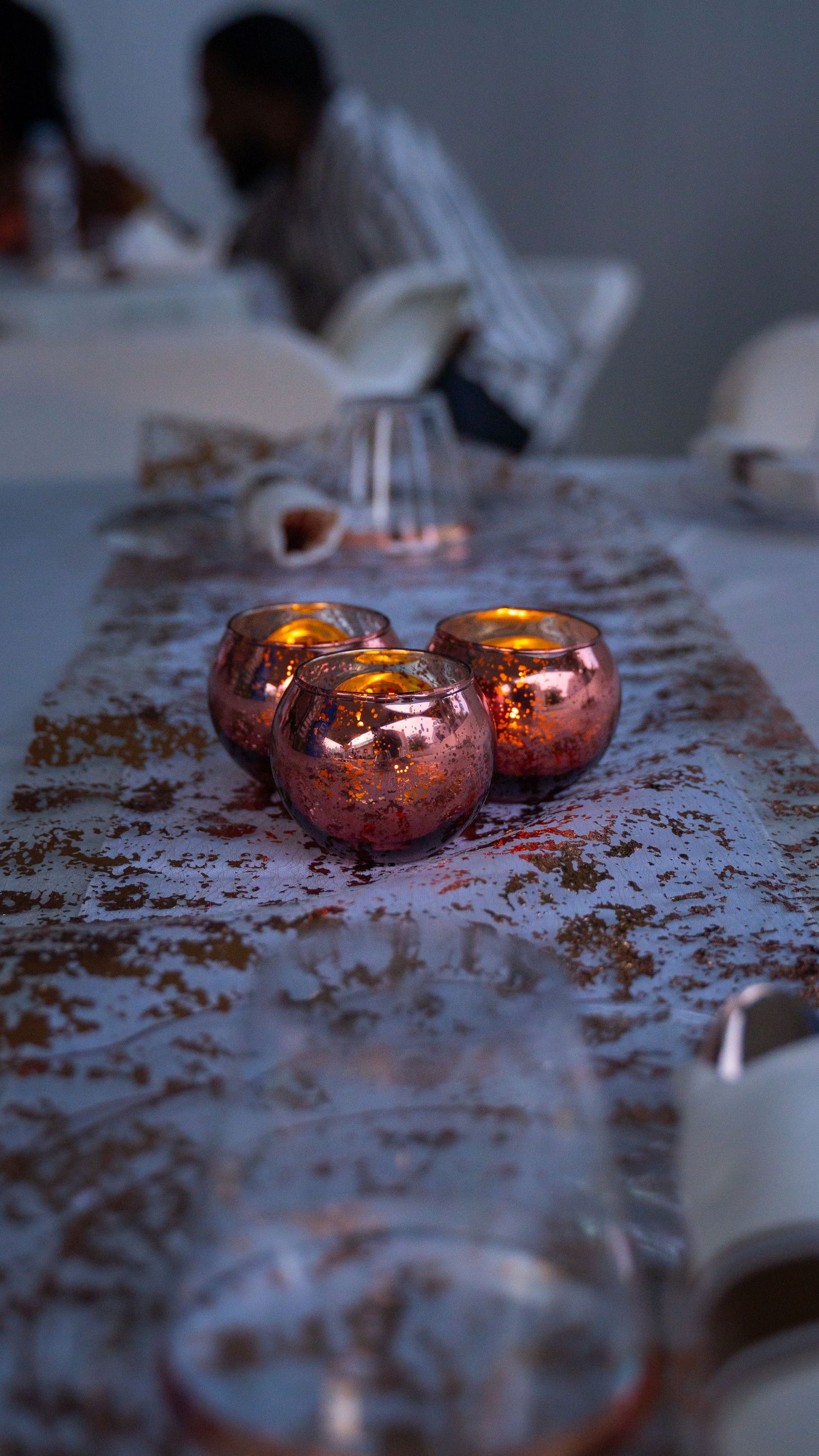 Three glowing rose gold mercury glass votives create warm ambiance on a textured table runner in dim lighting.