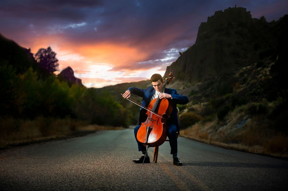 Musician playing cello on empty road during dramatic sunset with mountain silhouette.