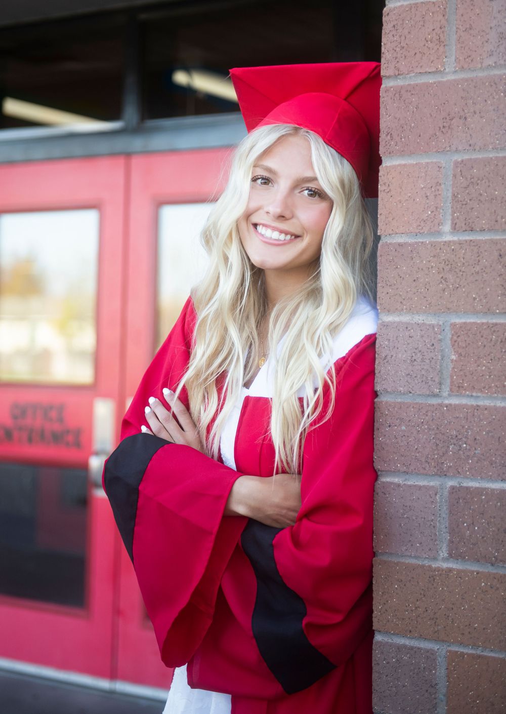 A graduate in a bright red cap and gown poses against a brick wall with long blonde hair and a cheerful smile.