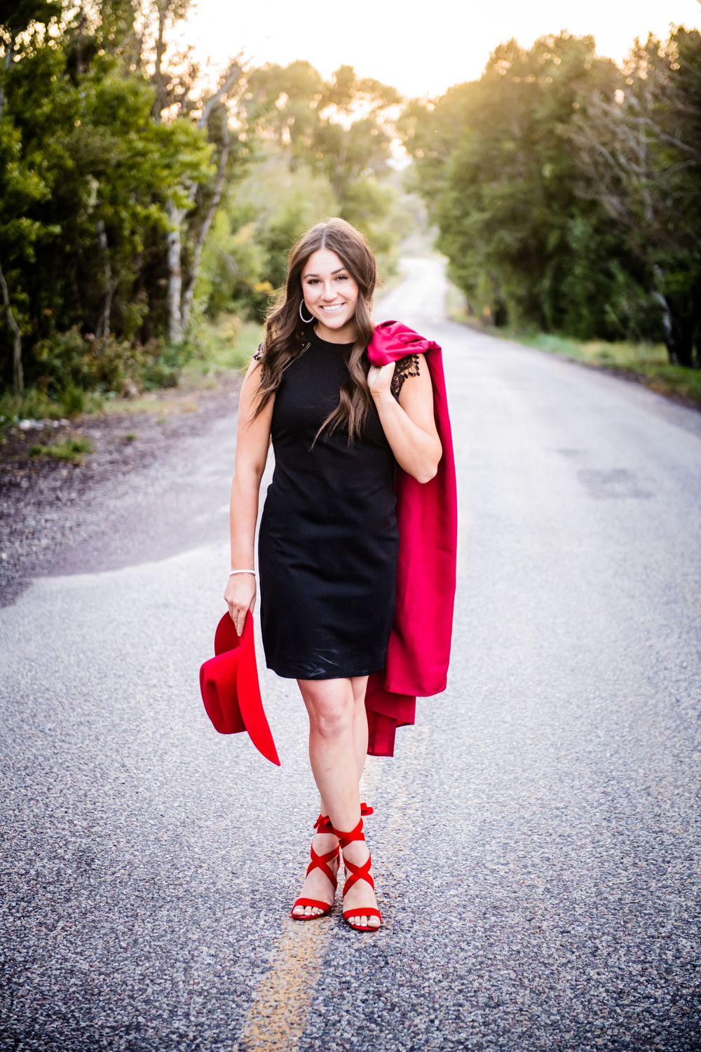 Fashion portrait on rural road featuring black dress and red accessories at sunset.