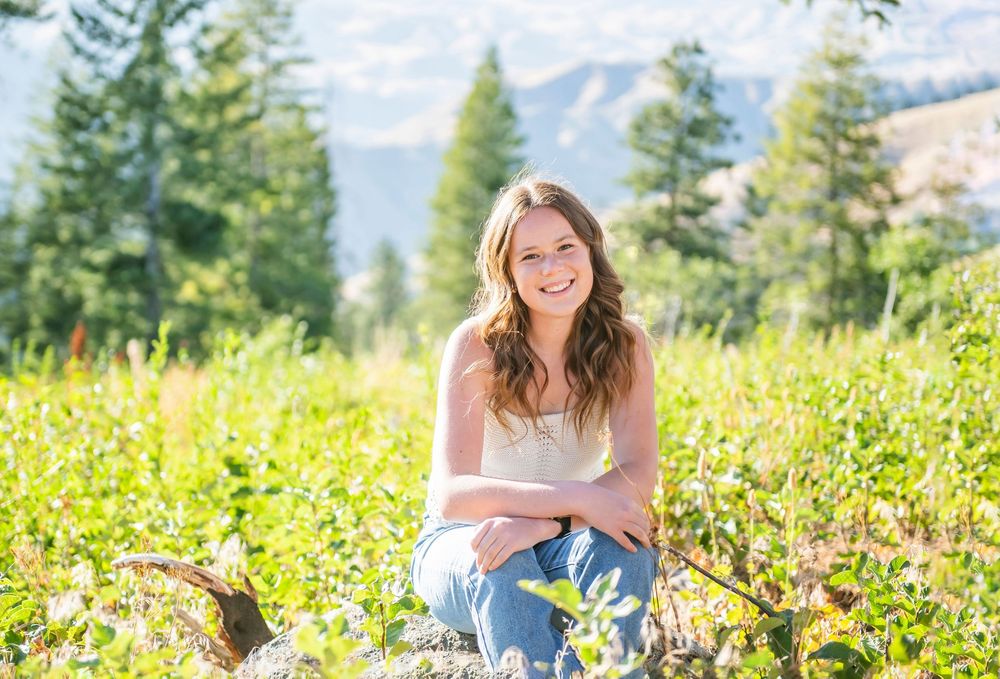 Someone sits in a mountain meadow surrounded by wildflowers and evergreen trees on a sunny summer afternoon.