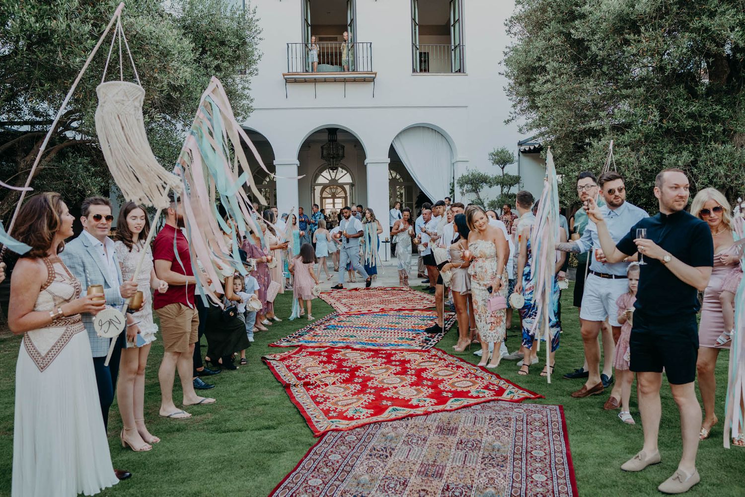 Guests gather around a red carpet aisle lined with vintage rugs at an outdoor garden ceremony.