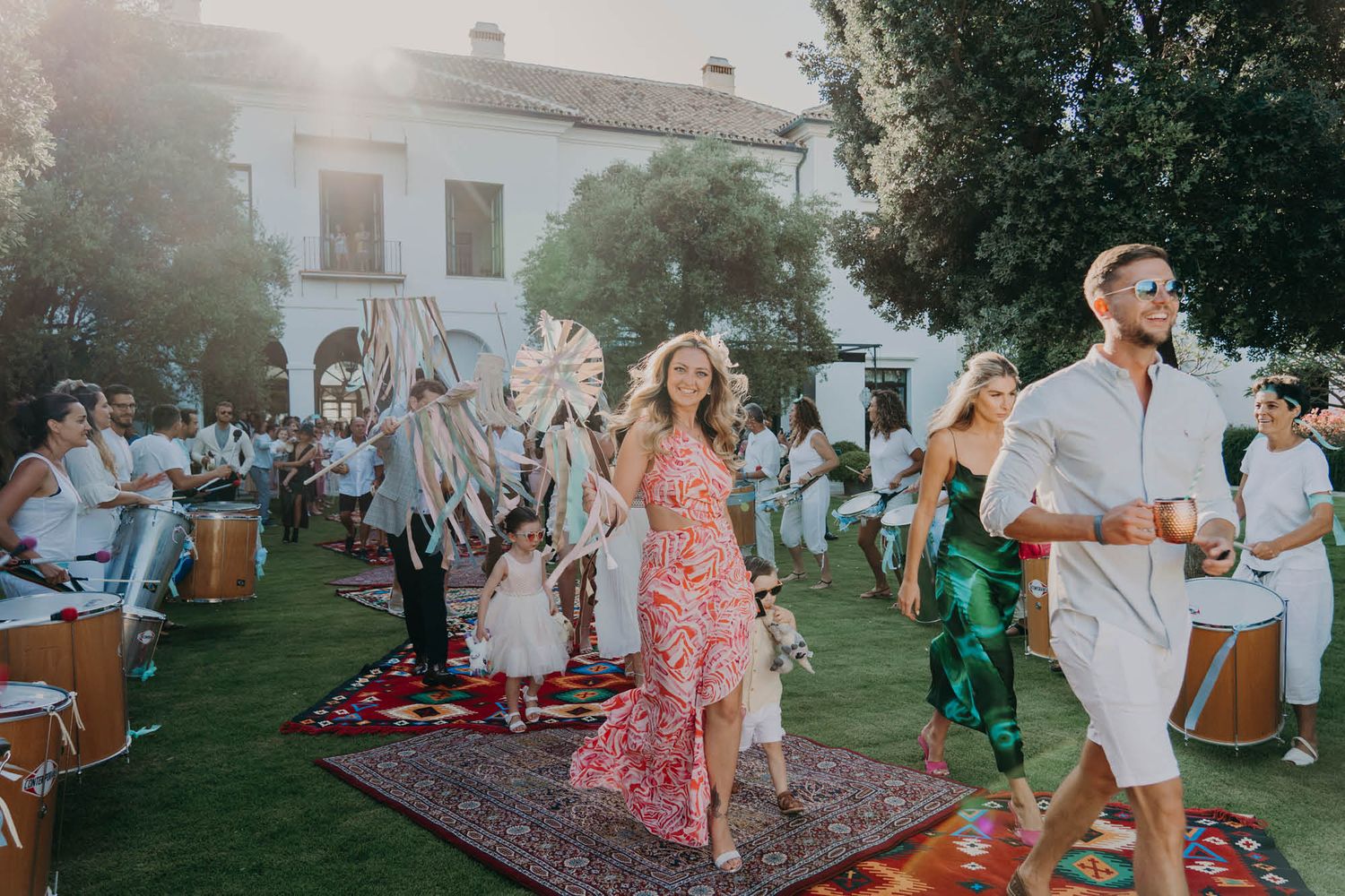 People dance and celebrate at an outdoor garden party with vintage rugs and decorations during a sunny afternoon.