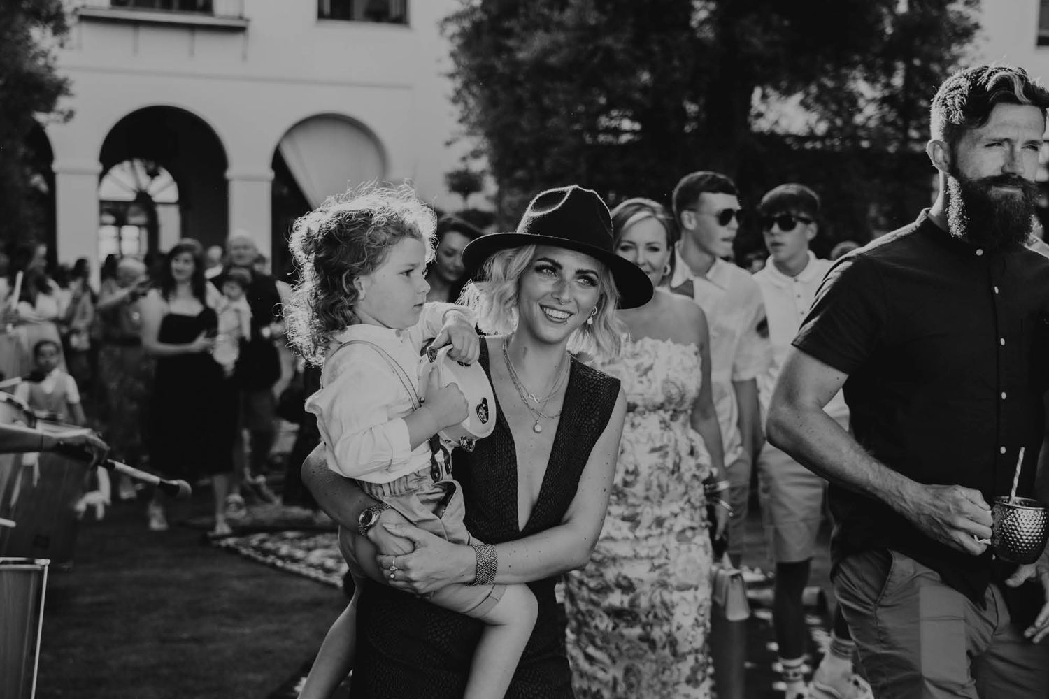 A black and white photo captures a happy outdoor crowd gathering at an event with arched architecture in the background.