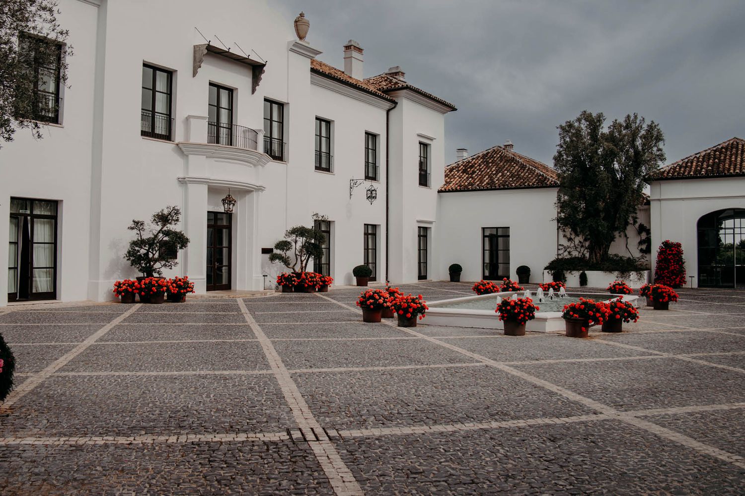 White colonial style buildings with terracotta roofs surround a stone courtyard adorned with red decorative planters.