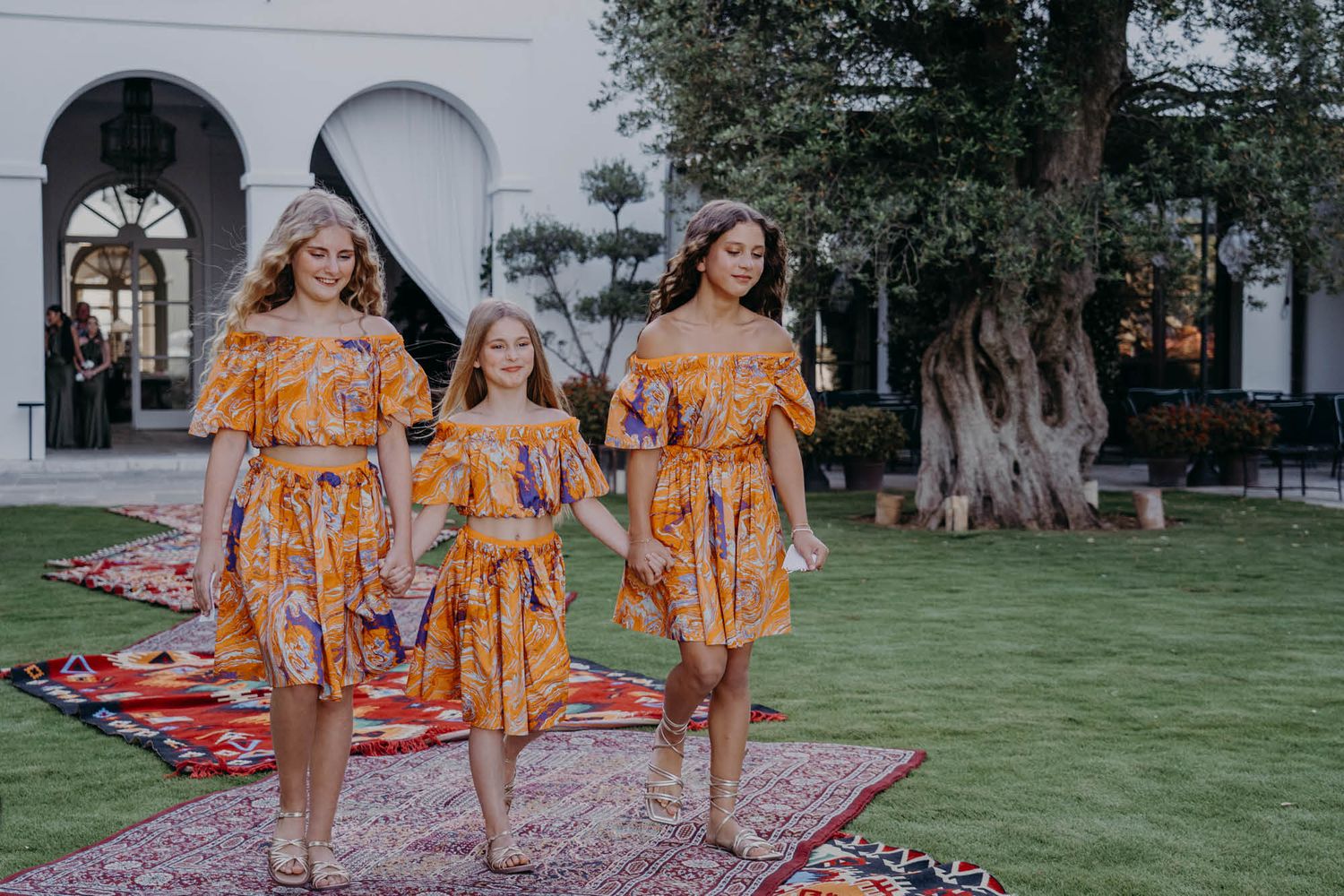 Three people wearing matching yellow floral summer dresses walk together on a lawn with decorative rugs and arched doorways.