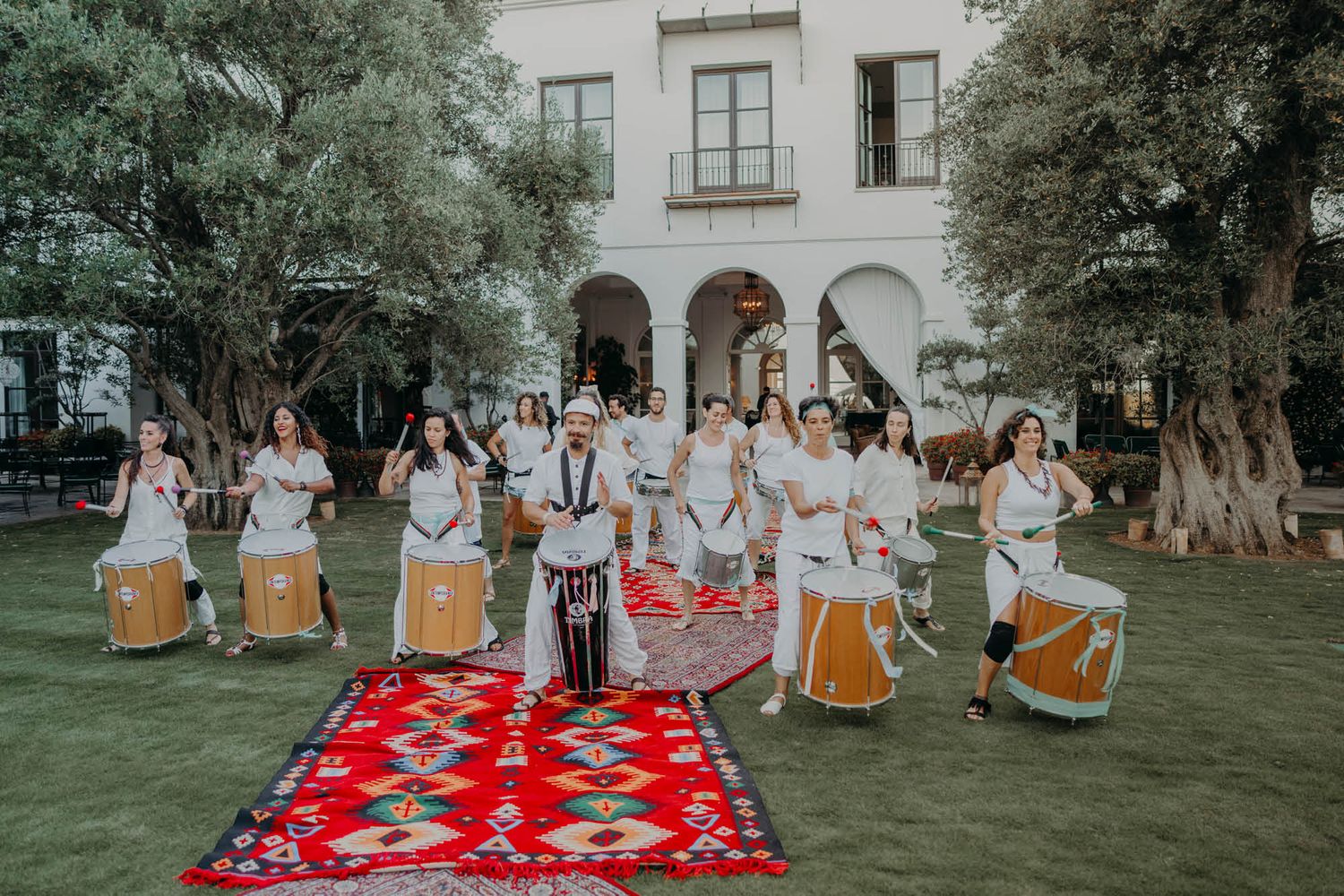 People in white dresses perform a traditional ceremony on a red carpet surrounded by trees and stone architecture.