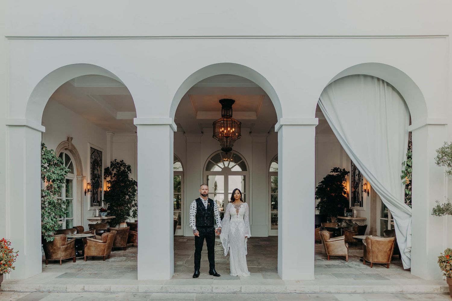 An elegant white archway frames a couple standing under a chandelier at a luxury wedding venue.