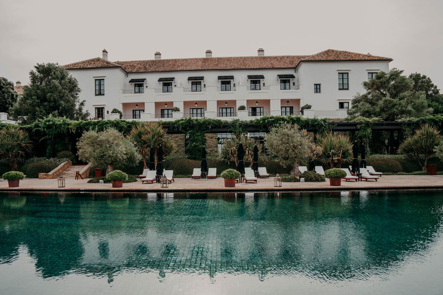 A luxurious white Mediterranean-style hotel building with a pristine turquoise swimming pool and lounge chairs.