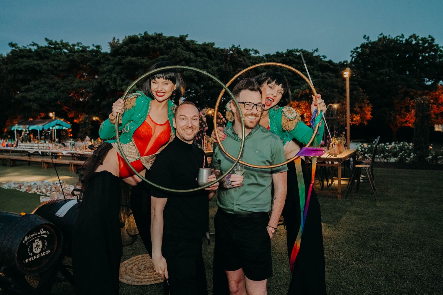 Performers with golden hula hoops pose together at an outdoor evening event with trees in the background.