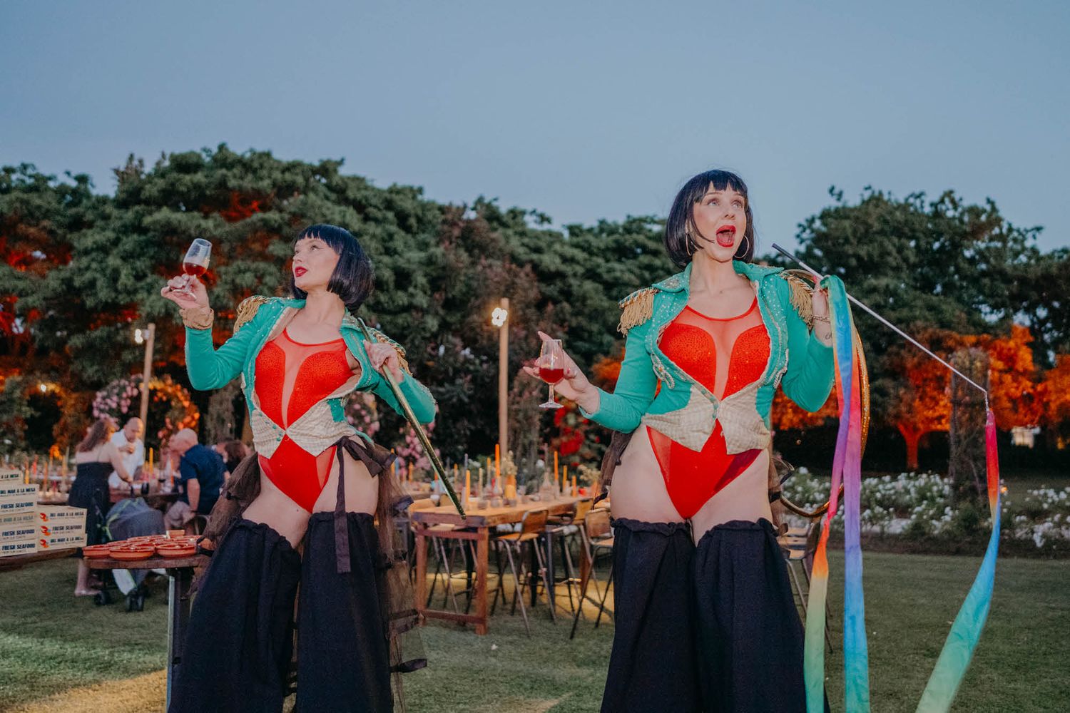 Two performers in matching costumes with hula hoops entertain guests at an outdoor garden venue at dusk.