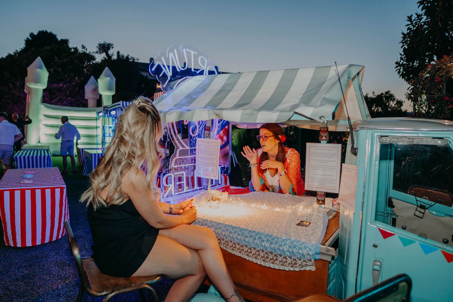 A vintage turquoise food truck serves customers at an outdoor evening event with colorful lighting and striped awning.