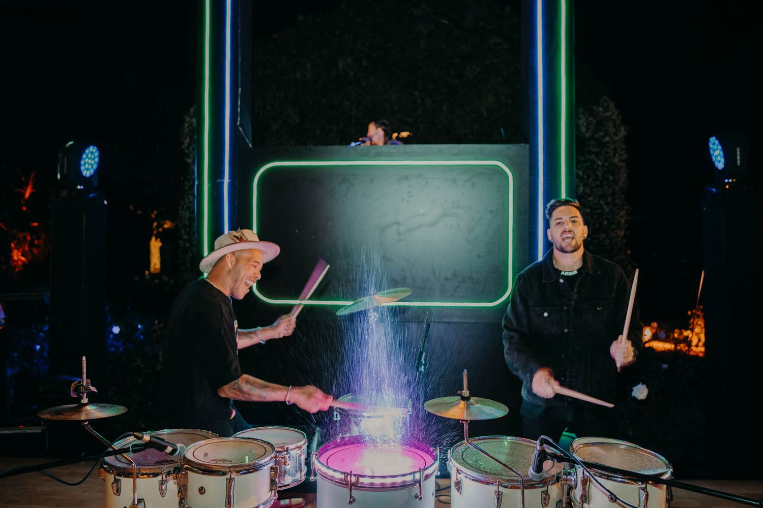 Two drummers perform with glowing drumsticks on a dark stage with colorful lighting effects.