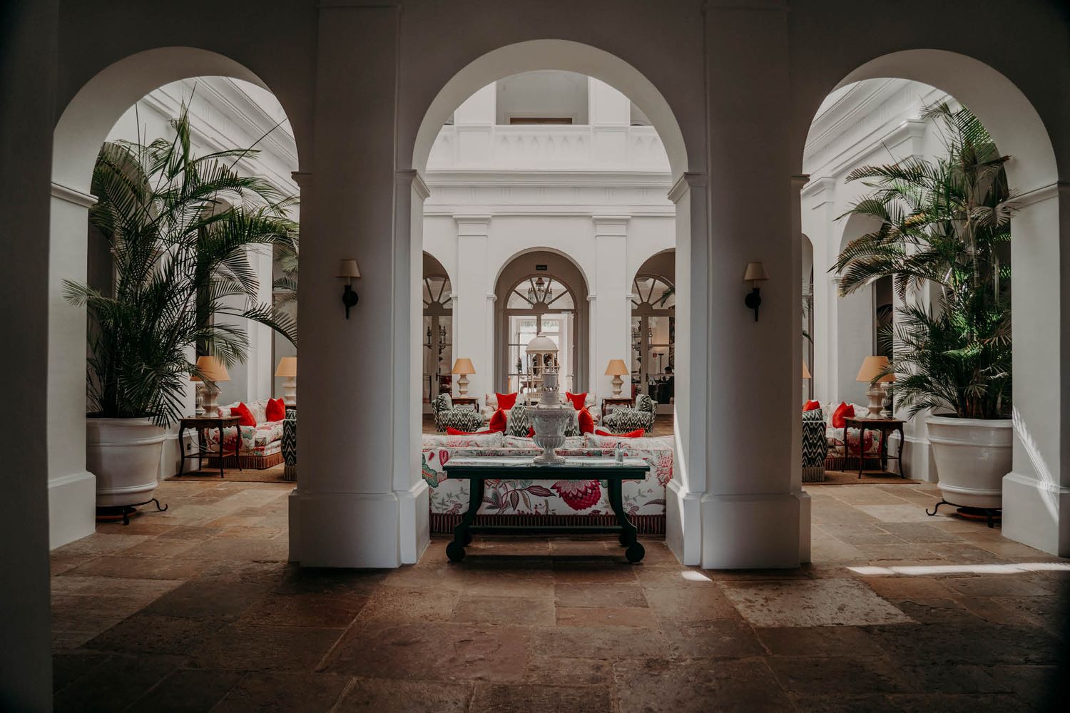 An elegant white arched hallway with potted palms and red decor leading to a dining area.