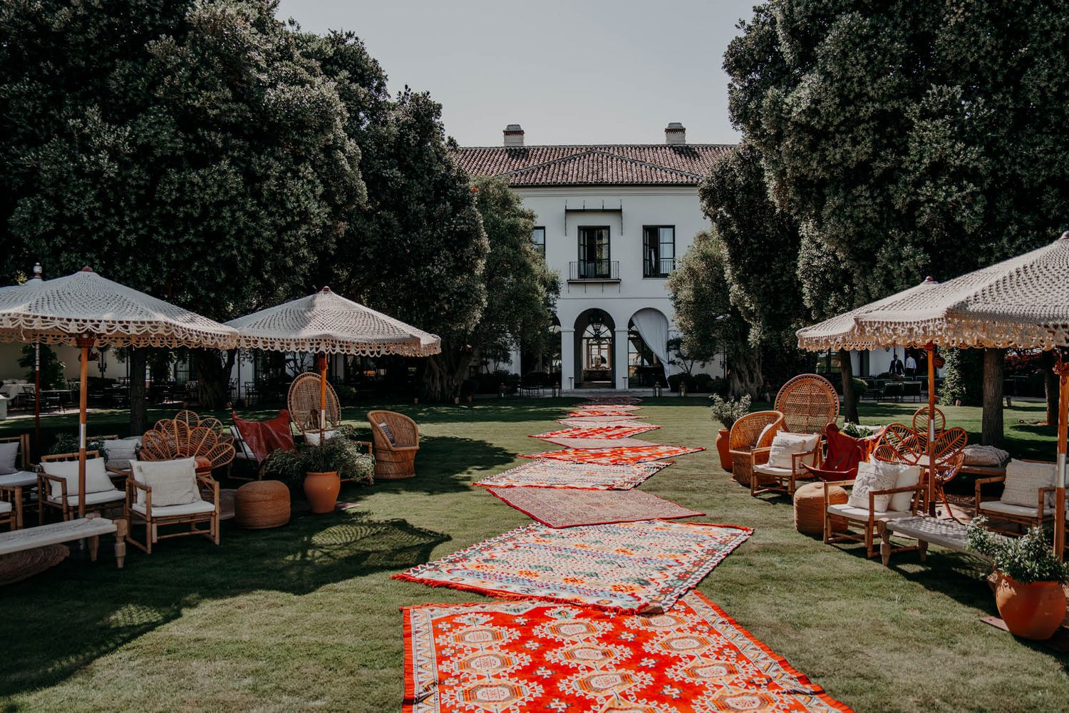 A romantic garden wedding setup with vintage rugs lining the aisle leading to a white mansion between tall trees.