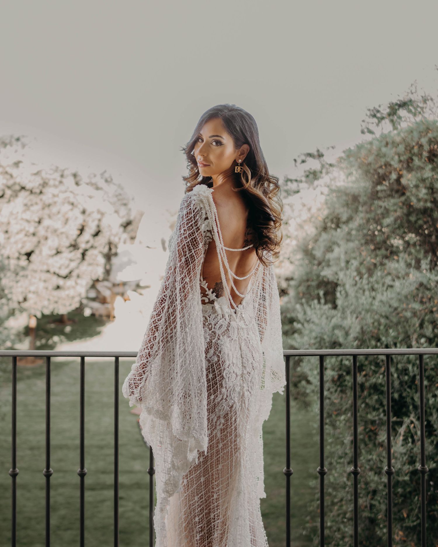 A bride in a delicate lace gown with flowing sleeves poses on a balcony overlooking a garden landscape.